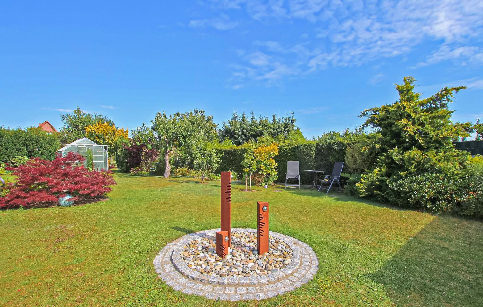 Un jardin agrémenté d’un élément décoratif circulaire en pierre, d’une serre et de divers arbres et arbustes.