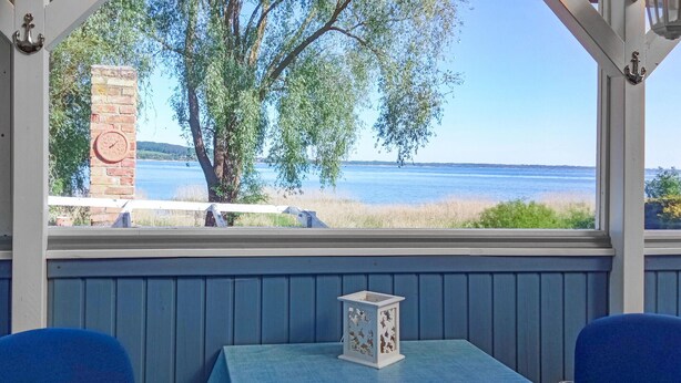 A table with a blue tablecloth, two blue chairs, a white lantern, and a view of a lake with trees and a brick wall.