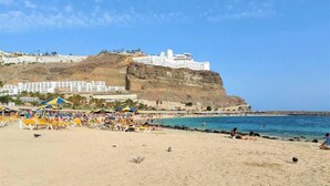Uma praia com espreguiçadeiras e guarda-sóis, um penhasco rochoso e um edifício no topo.