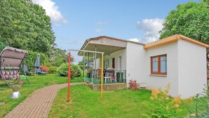 Une petite maison de plain-pied avec un porche, une balançoire et un jardin où poussent différentes plantes et fleurs.