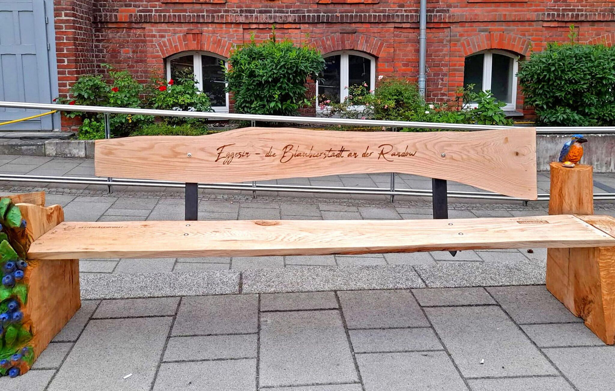 Een houten bankje met een gebeeldhouwde inscriptie in een vreemde taal, voor een bakstenen gebouw met ramen en beplanting.