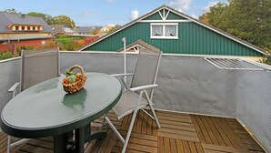 Um deck de metal verde com uma mesa redonda e duas cadeiras, uma cesta de vime com vegetais, e um edifício verde ao fundo.