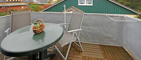 En grøn metalterrasse med et rundt bord og to stole, en fletkurv med grøntsager, og en grøn bygning i baggrunden.