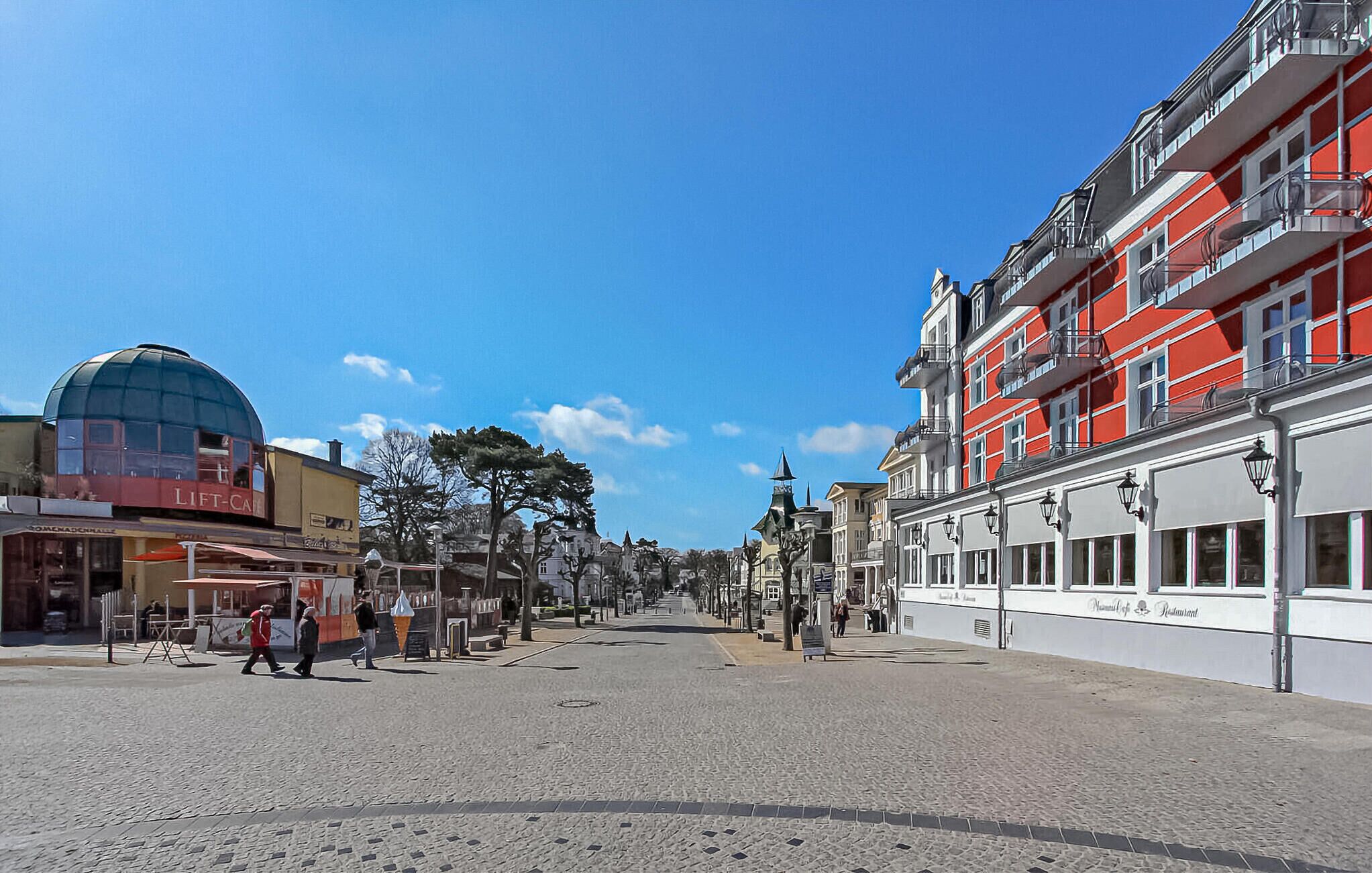 Une rue pavée bordée de bâtiments historiques, dont un immeuble rouge et blanc doté de balcons et un immeuble jaune surmonté d’un dôme.