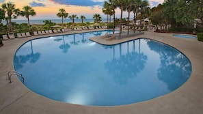 A large, curved swimming pool with lounge chairs and palm trees, surrounded by a landscaped area with a view of the ocean. - 𝐅𝐢𝐧𝐝𝐢𝐧𝐠 𝐌𝐞𝐢𝐥𝐢:Beachfront Fun, Grande Ocean Resort+Amenities Included (Hilton Head Island)