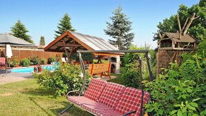A patio area with a red plaid bench, a wooden swing, a stone fireplace, and a pool in the background. - Cozy home in Kummerow with sauna (Kummerow)