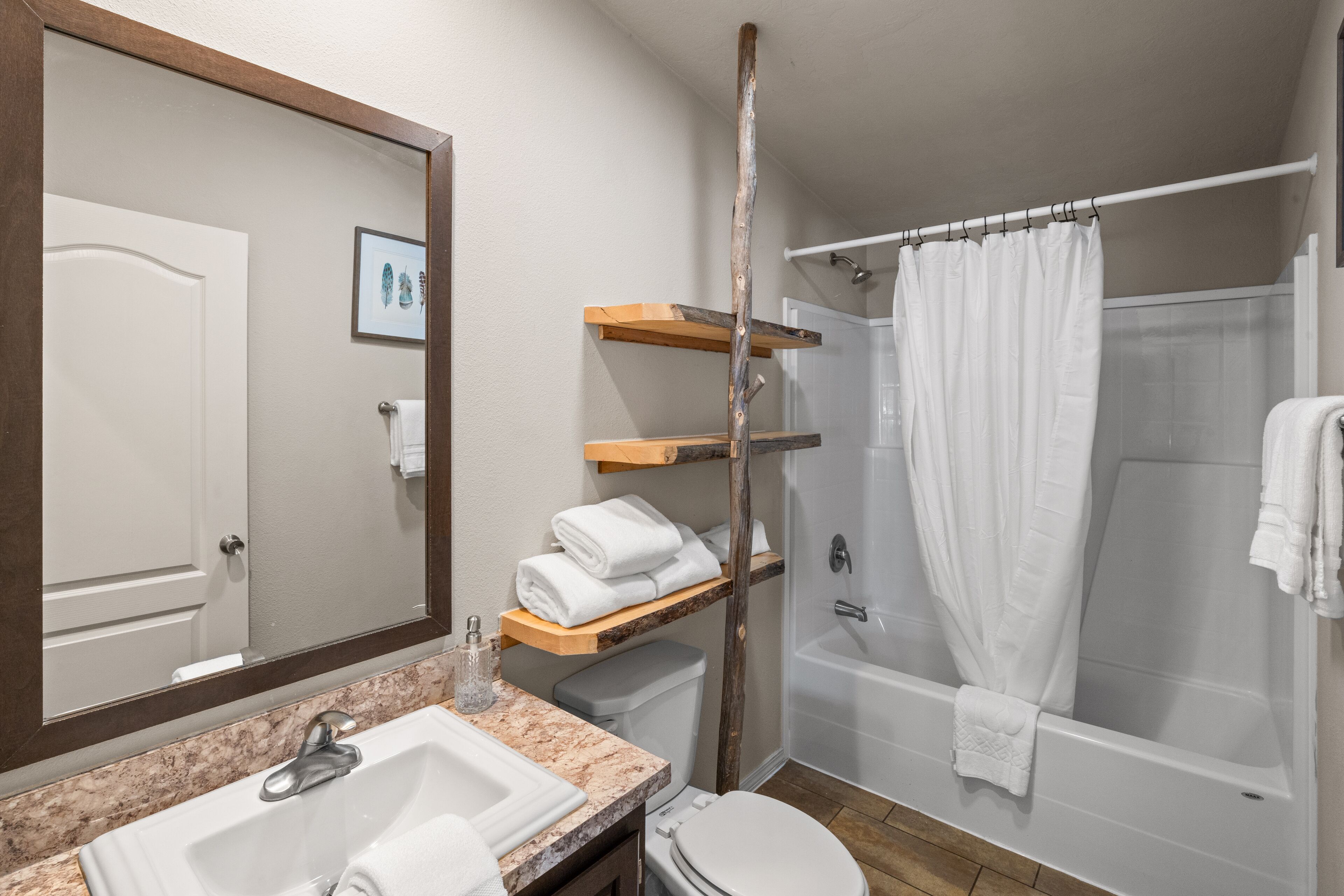 Un baño con lavamanos, inodoro y ducha. Hay repisas de madera sobre el inodoro y un espejo grande sobre el lavamanos.