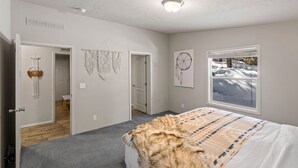 A bedroom with a large bed, a window with a view of a snowy landscape, a door leading to another room, and wall decorations. - Dreamcatcher (West Yellowstone)