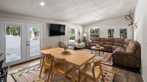 A living room with a wooden dining table, brown leather sofa, and a TV mounted on the wall. - Dreamcatcher (West Yellowstone)