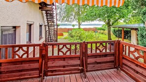 Une terrasse en bois avec une rambarde, un espace abrité doté d’un auvent à rayures, et une vue sur un jardin et un lac.