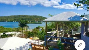 A covered outdoor seating area with tables and chairs, overlooking a body of water and a hilly landscape. - 2bdrm, Water deck views Bag Drop
 (Culebra)