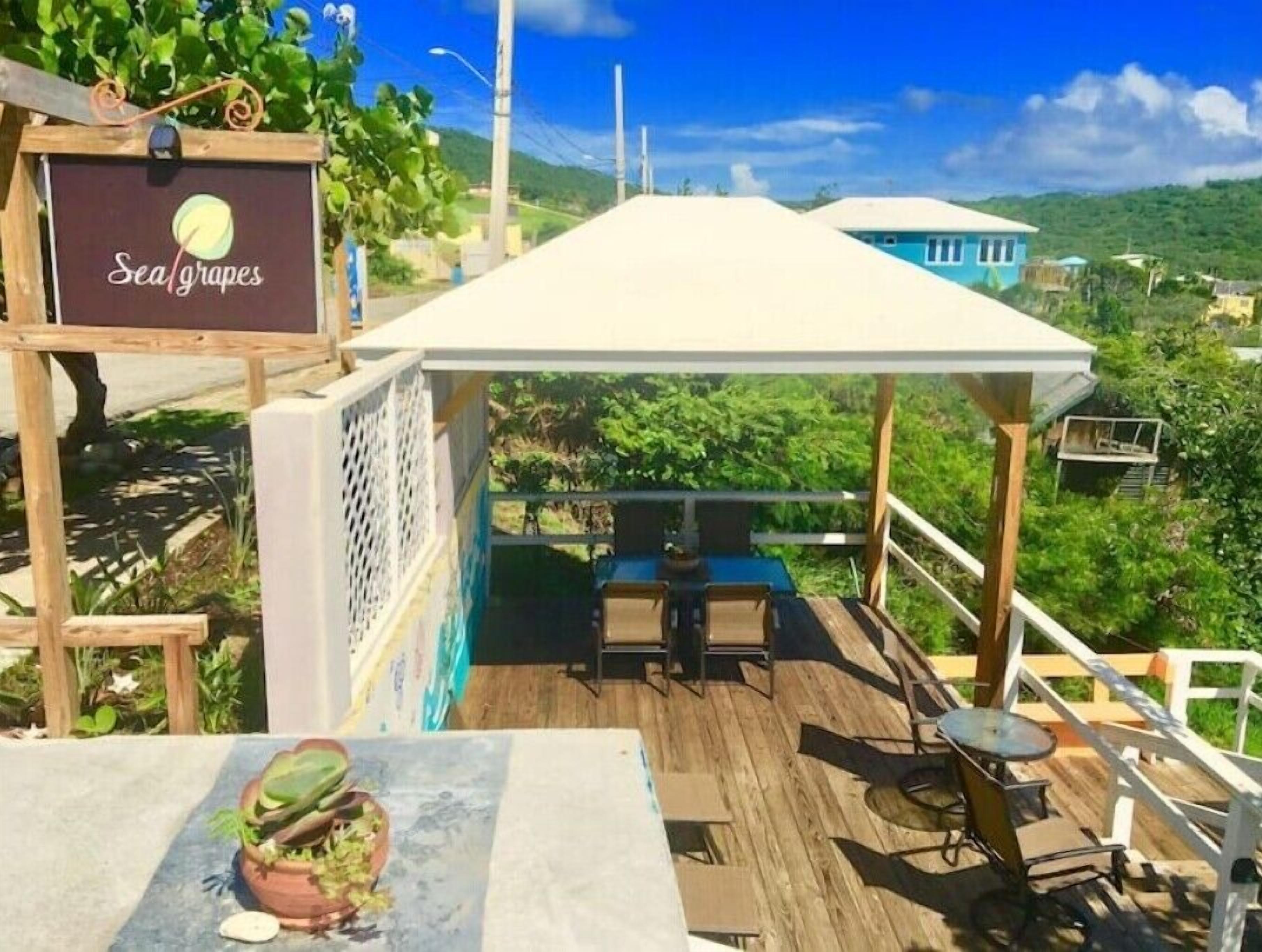 A wooden deck with a table and chairs, a sign reading "Sea grapes", and a view of a lush green landscape.