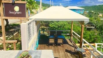 A wooden deck with a table and chairs, a sign reading "Sea grapes", and a view of a lush green landscape.