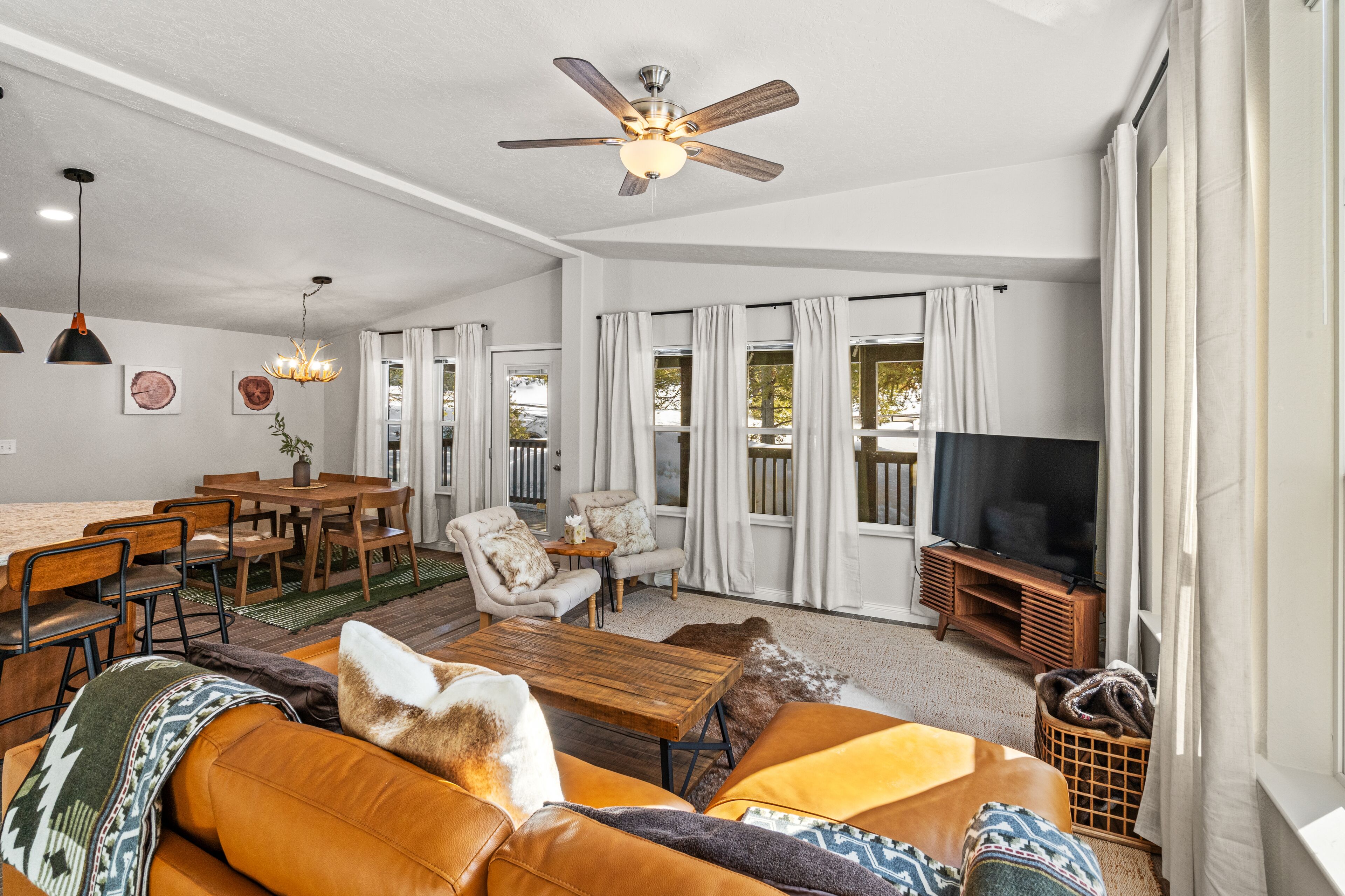 A spacious living room with a large sofa, a wooden coffee table, and a ceiling fan. There is a dining area with a table and chairs, and a television on a stand.