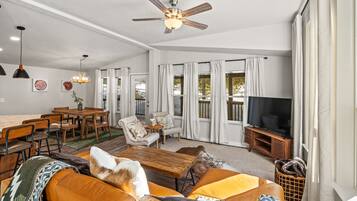A spacious living room with a large sofa, a wooden coffee table, and a ceiling fan. There is a dining area with a table and chairs, and a television on a stand.