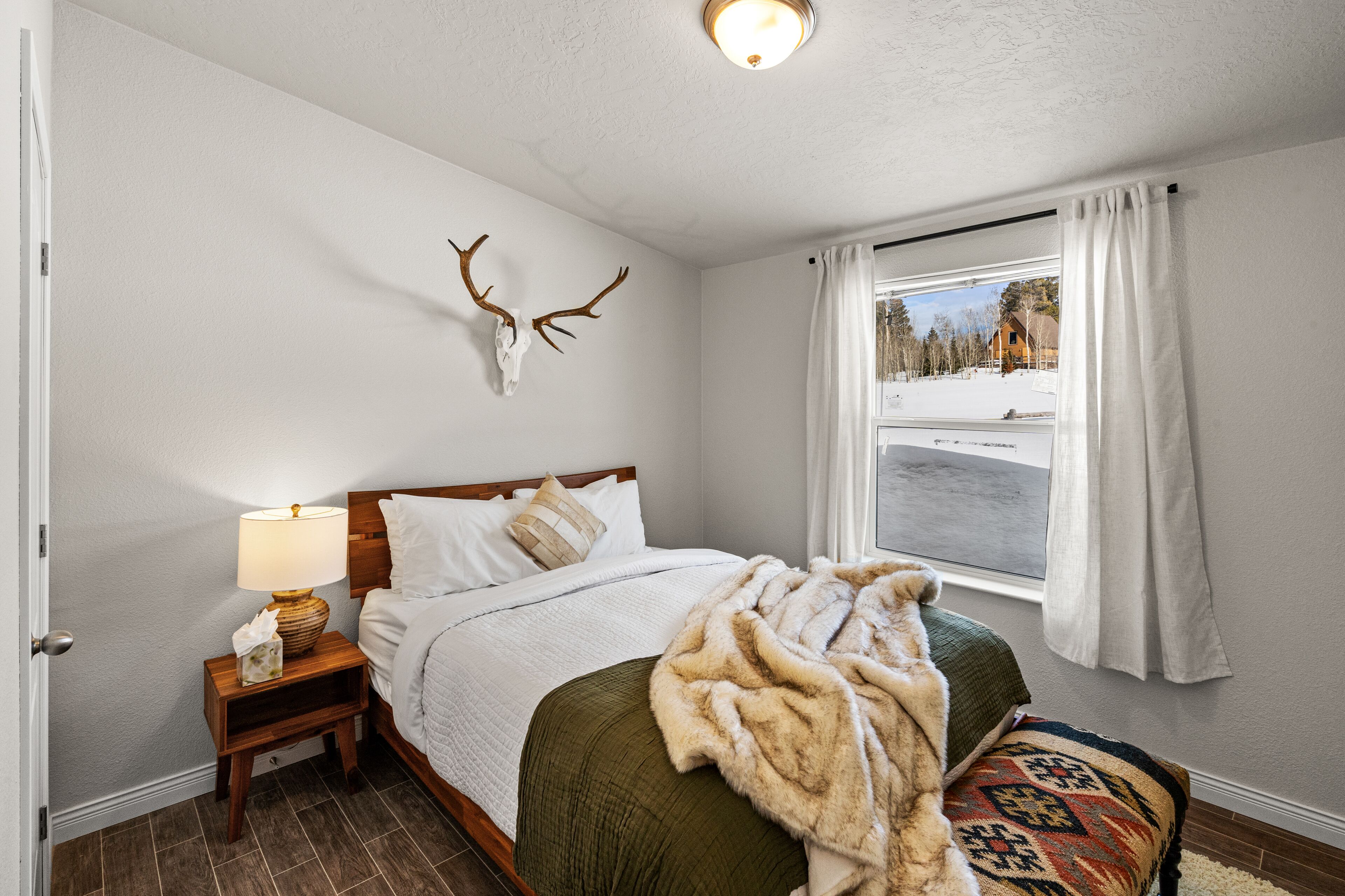 Un dormitorio con cama, mesita de noche, lámpara, manta, decoración de cráneo de venado en la pared, ventana con cortinas y vista a un paisaje nevado.
