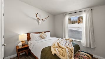 Un dormitorio con cama, mesita de noche, lámpara, manta, decoración de cráneo de venado en la pared, ventana con cortinas y vista a un paisaje nevado.