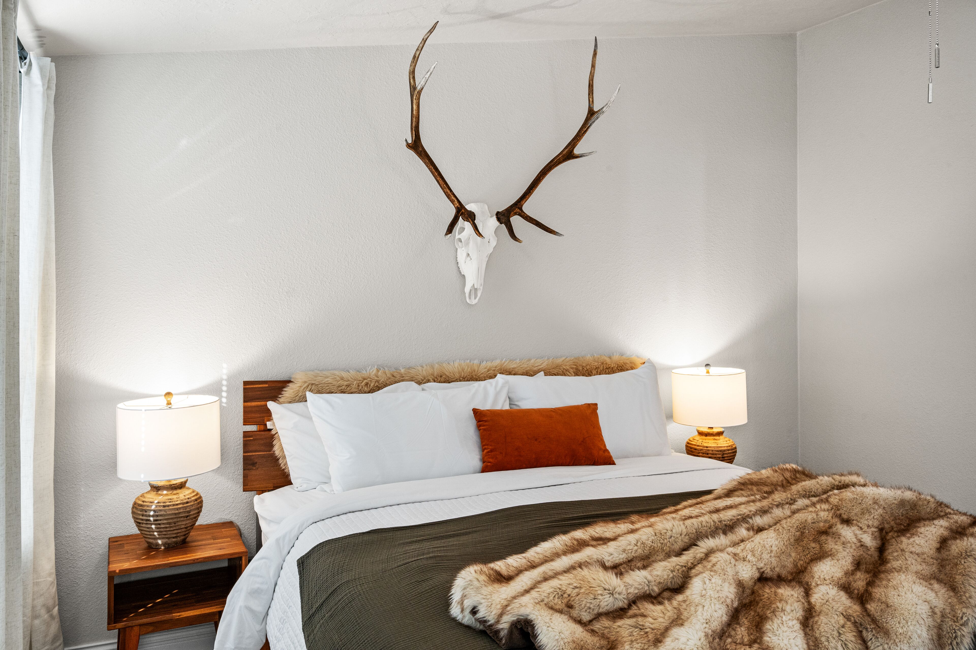 A bedroom with a bed, two bedside lamps, a fur throw, and a deer skull mounted on the wall.