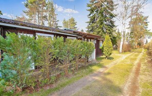 Un chalet rustique avec une façade en bois, un chemin de gravier menant à l’entrée et une végétation luxuriante qui entoure les lieux.
