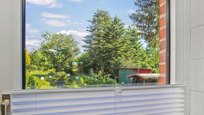 Une fenêtre donnant sur un jardin, un bâtiment rouge et un ciel dégagé.