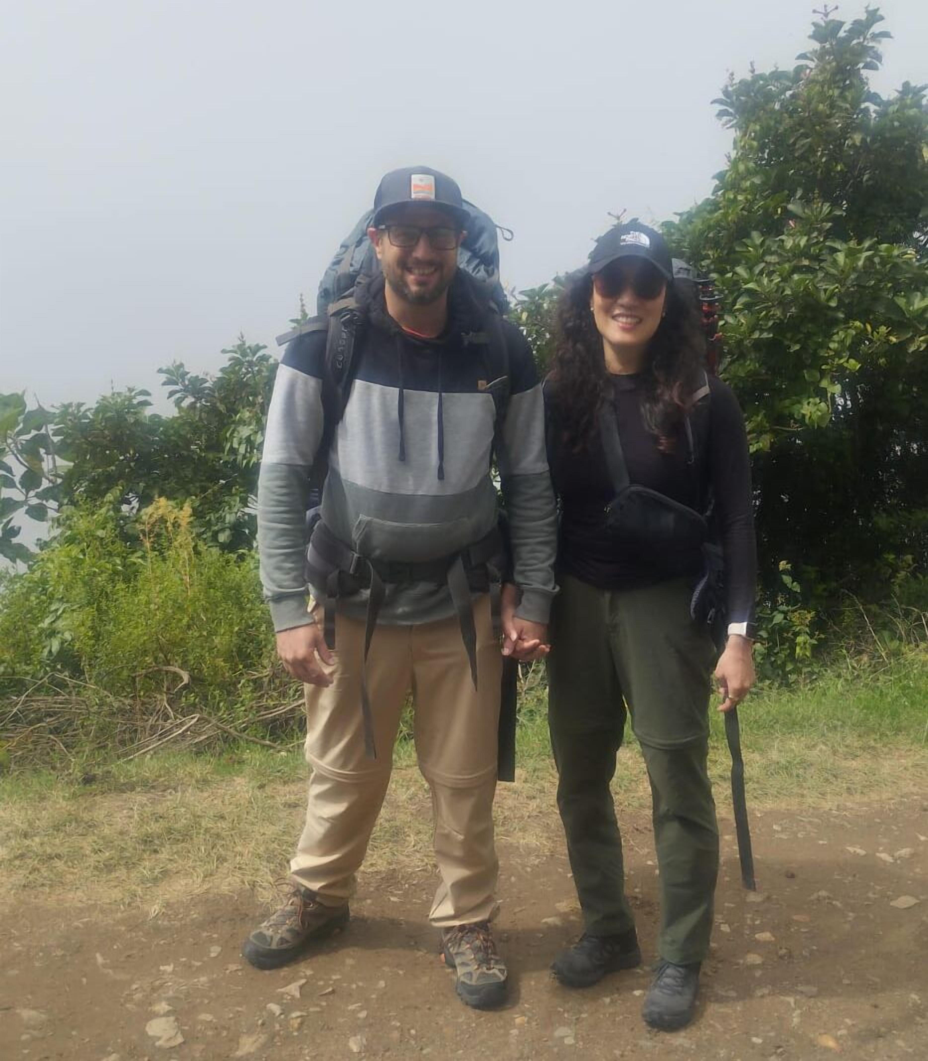 Two people standing on a dirt path with backpacks, surrounded by greenery.