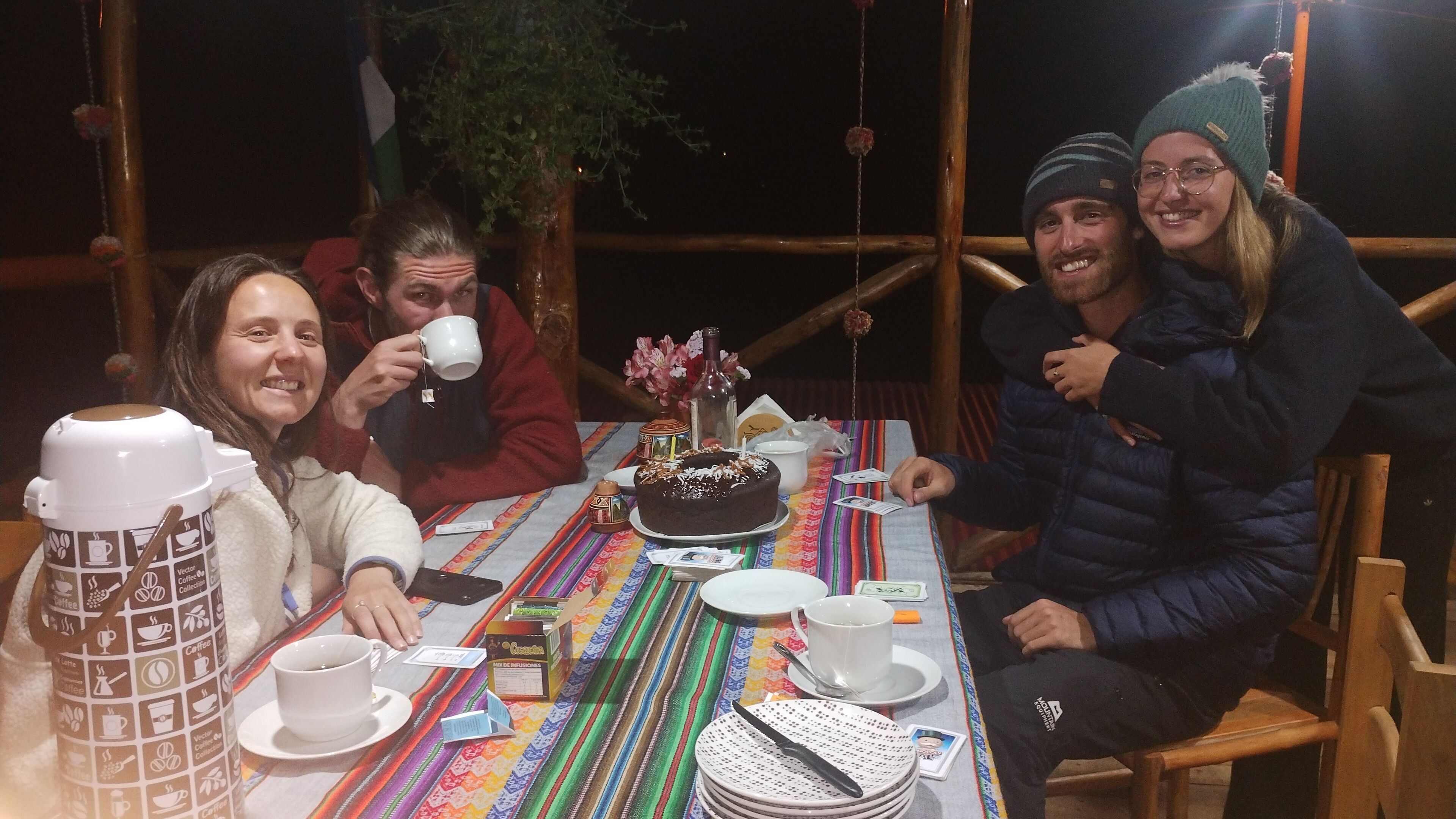 A group of people sitting around a table with a cake, coffee cups, and a bottle.
