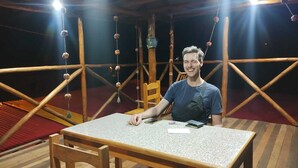 A person seated at a table in a room with a wooden floor and a red carpet. - the wooden house choquequirao (San Pedro de Cachora)