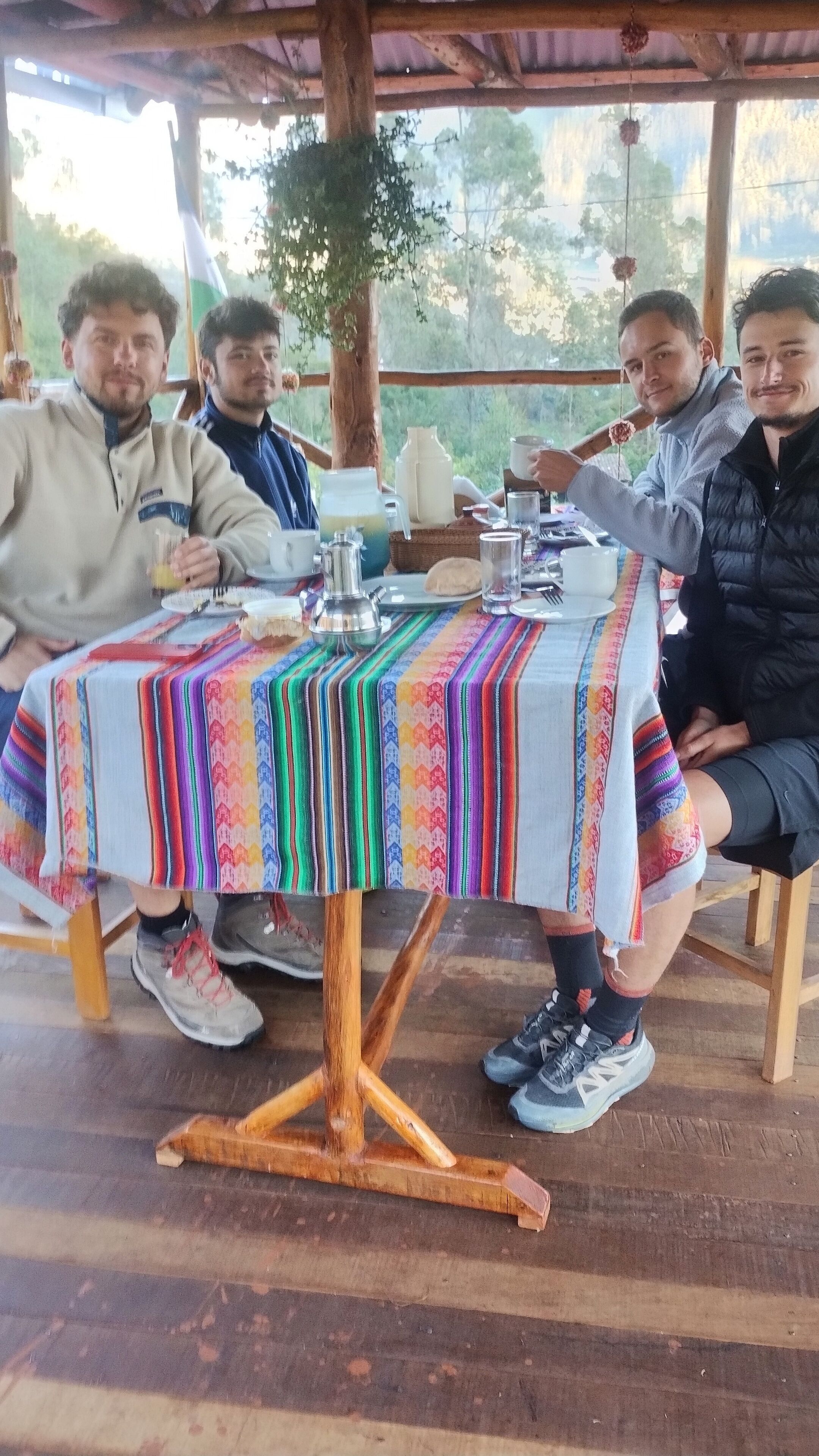 Eine Gruppe von Personen sitzt an einem Holztisch mit einer bunten Tischdecke, unter einer hölzernen Überdachung.