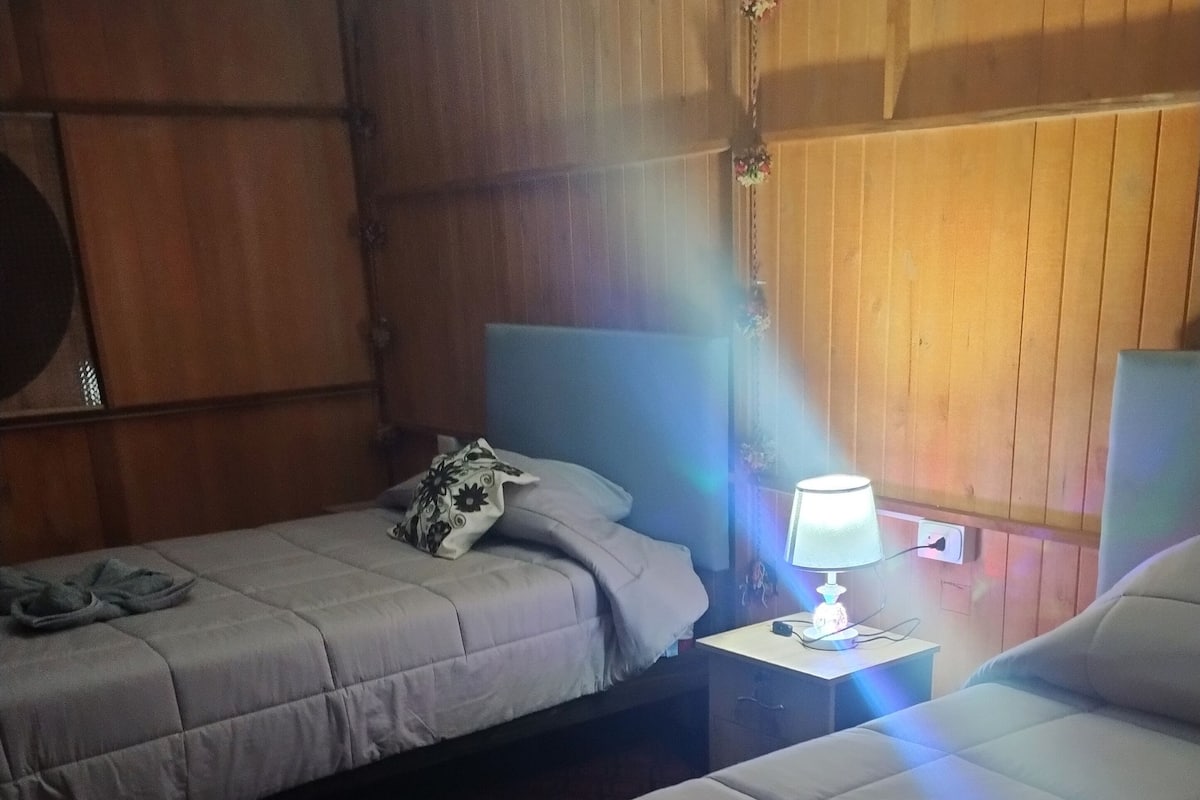 Habitación de hotel con dos camas, una mesita de noche con lámpara y paneles de madera en las paredes.
