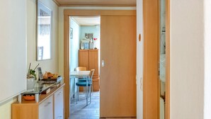 Ein schmaler Flur mit Küchenzeile links, inklusive Schränken und einer Arbeitsplatte mit Schüssel und Pflanze. Im Hintergrund ist ein Esstisch mit Stühlen zu sehen.