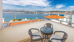 A balcony with two wicker chairs and a small table, overlooking a coastal town and a large body of water.