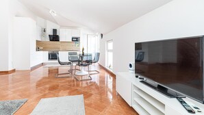 A modern living room with a flat-screen TV, a dining area with a glass table and chairs, and a kitchen with white cabinets and a wooden countertop.