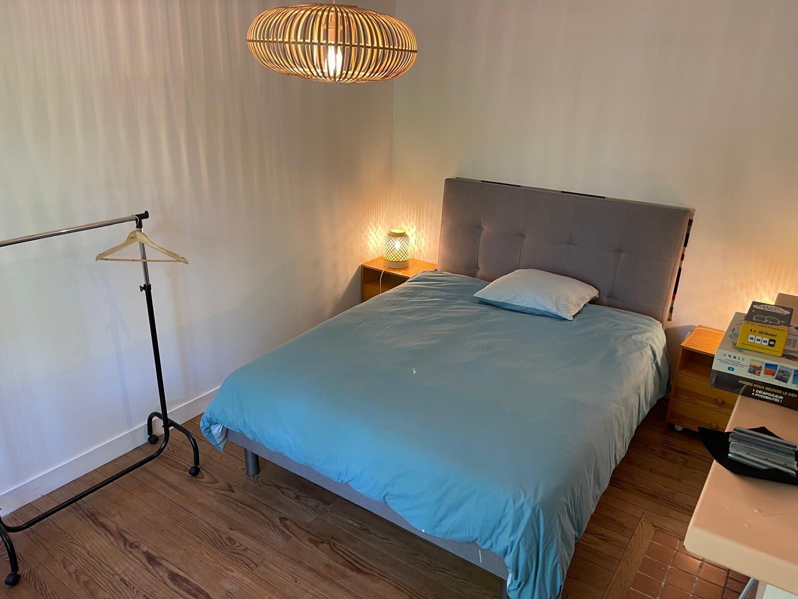A bedroom with a bed, a hanging light fixture, a bedside table with a lamp, and a wooden floor.