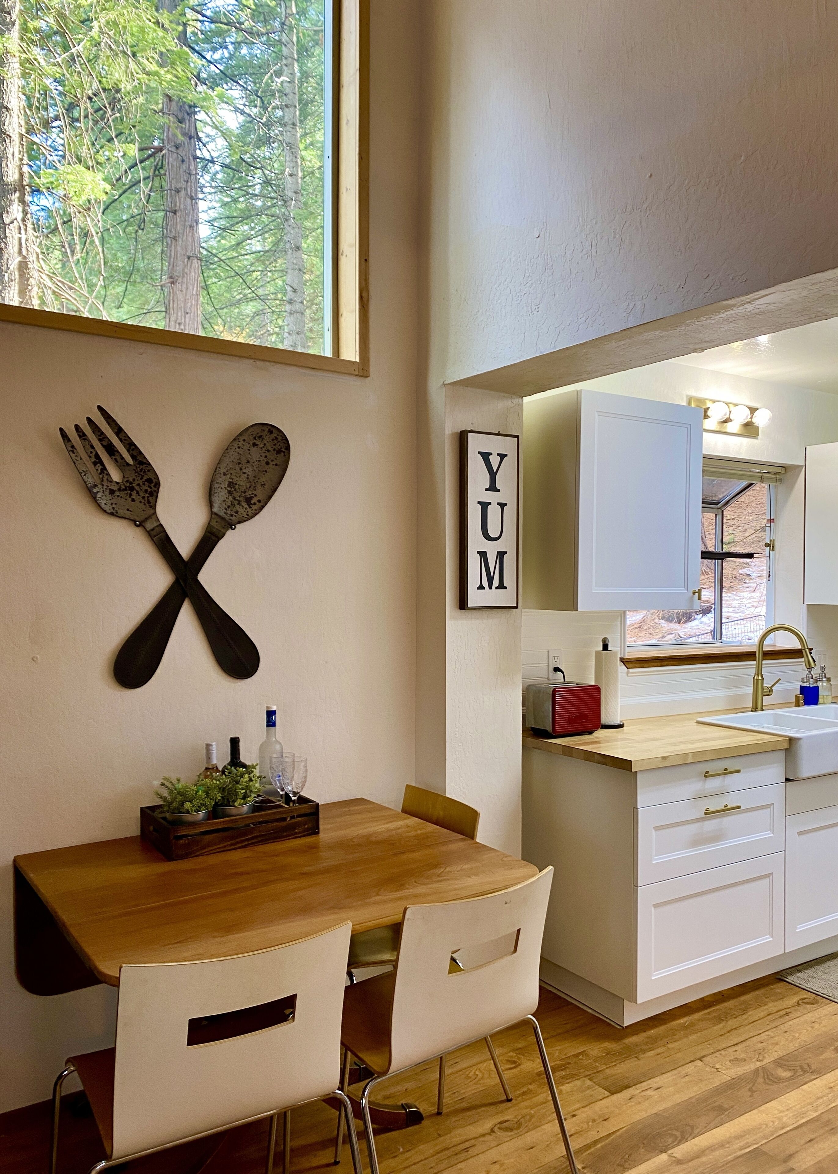 A modern kitchen with white cabinets, a wooden dining table, and a window overlooking greenery.