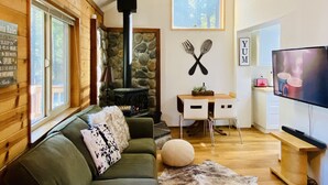 A cozy cabin interior with a stone fireplace, a wooden ceiling fan, a dining table with chairs, and a television.