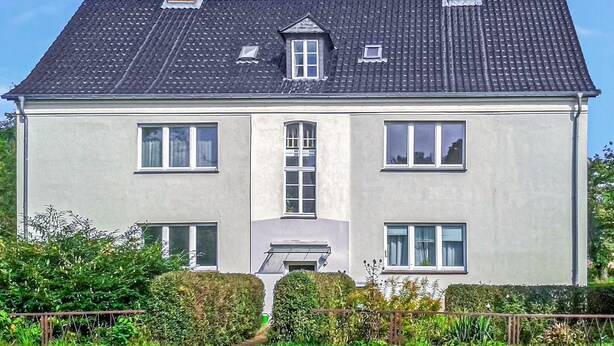 Une maison à deux étages, avec une façade grise, un toit recouvert de tuiles sombres et de nombreuses fenêtres.