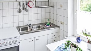 Une cuisine avec des meubles blancs, un évier à deux bacs et une cuisinière. On y voit des ustensiles de cuisine accrochés au mur, une fenêtre avec des plantes et une plante en pot posée sur le comptoir.