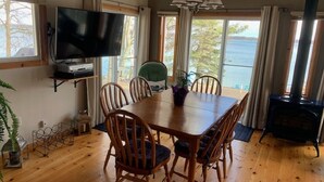 Uma sala de jantar com mesa e cadeiras de madeira, televisão numa prateleira e vista para o mar através de grandes janelas.