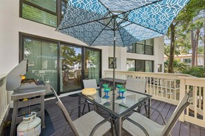 Une terrasse extérieure avec une table en verre, des chaises, un barbecue et un parasol à motifs.