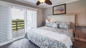 Ein Schlafzimmer mit einem großen Bett, einem Deckenventilator, Blick durch eine Schiebetür auf einen Balkon und einem gerahmten Bild an der Wand.