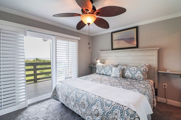 A bedroom with a large bed, a ceiling fan, a view through a sliding door to a balcony, and a framed picture on the wall.