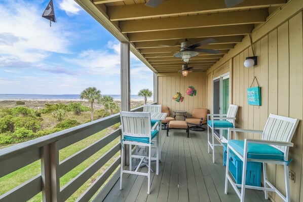 A covered outdoor seating area with a view of the beach, featuring white and blue chairs, a table, and a ceiling fan.