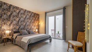 Une chambre moderne avec un lit, une chaise, une table de chevet, un mur orné d’un motif de feuilles et une porte-fenêtre coulissante donnant sur un balcon.