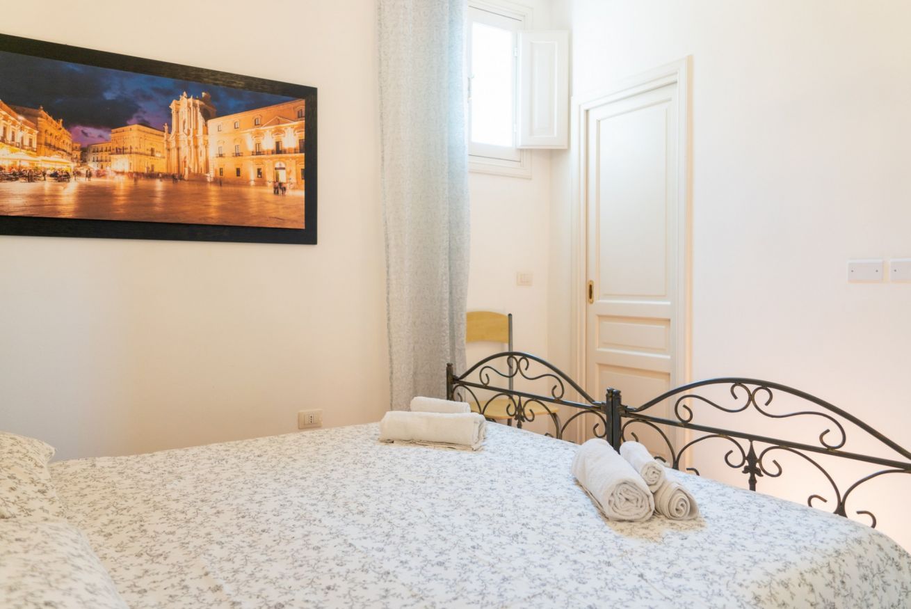 Une chambre d’hôtel avec un lit, un tableau représentant un paysage urbain, une fenêtre avec des rideaux et une porte.