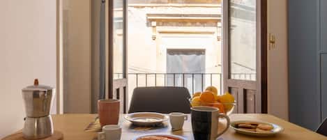 A breakfast table with a coffee maker, cups, a plate with a pastry, and a bowl of fruit.