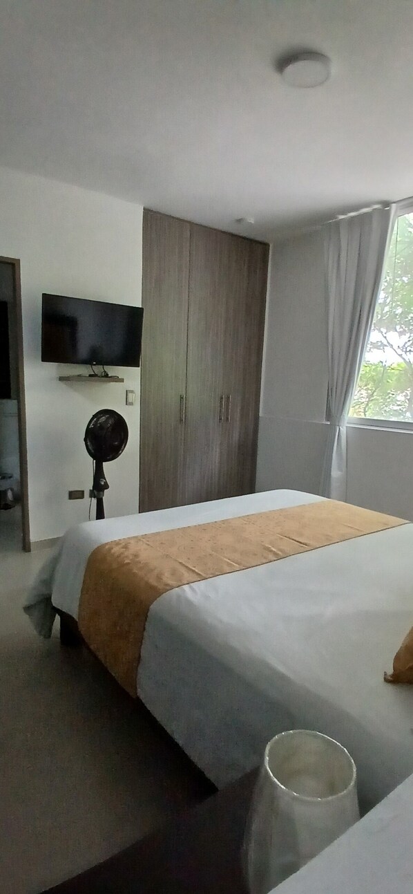 Una habitación de hotel con cama, televisión montada en la pared, ventilador y ventana con cortinas.