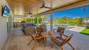A covered patio with a dining set, ceiling fans, and a view of a swimming pool and greenery.