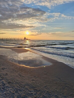 Una playa con un muelle que se adentra en el mar, olas suaves lamiendo la orilla y un atardecer al fondo.