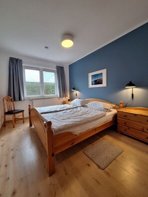 A bedroom with a wooden bed, a chair, a nightstand, a lamp, and a window with curtains. - Birkenstr. 14 Wg.2 "timeout Oasis" - Entspannung Pur: Timeout Oasis Ferienwohnung in Strandnah (Zingst)