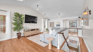 A spacious living area with a large grey sofa, a wooden cabinet, a TV mounted on the wall, and a staircase leading to an upper level. - Ocean View Haven - Walk to the Beach (Lincoln City)